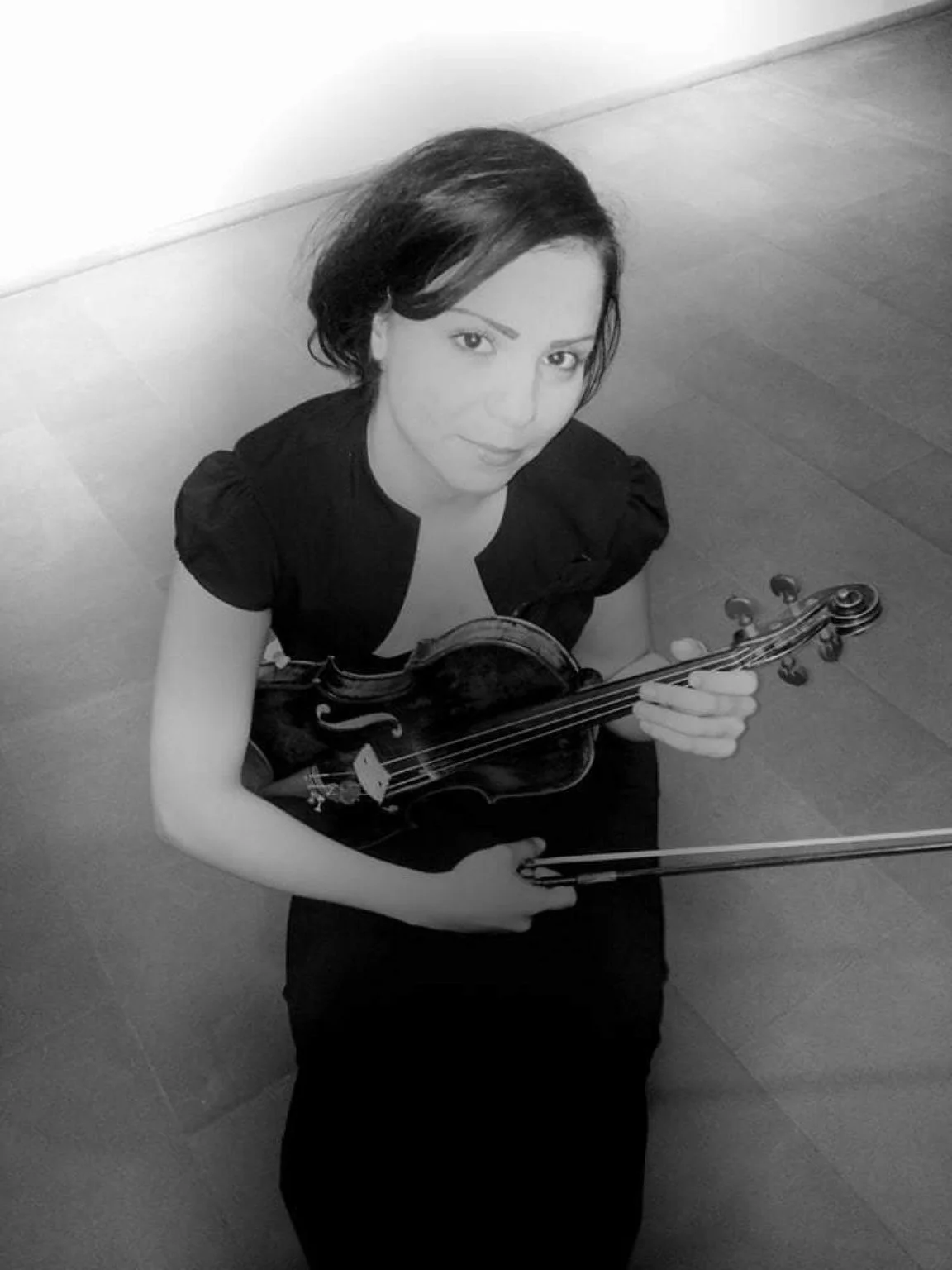 A woman sitting on the floor holding a violin and bow, looking up at the camera, in a black and white photo.