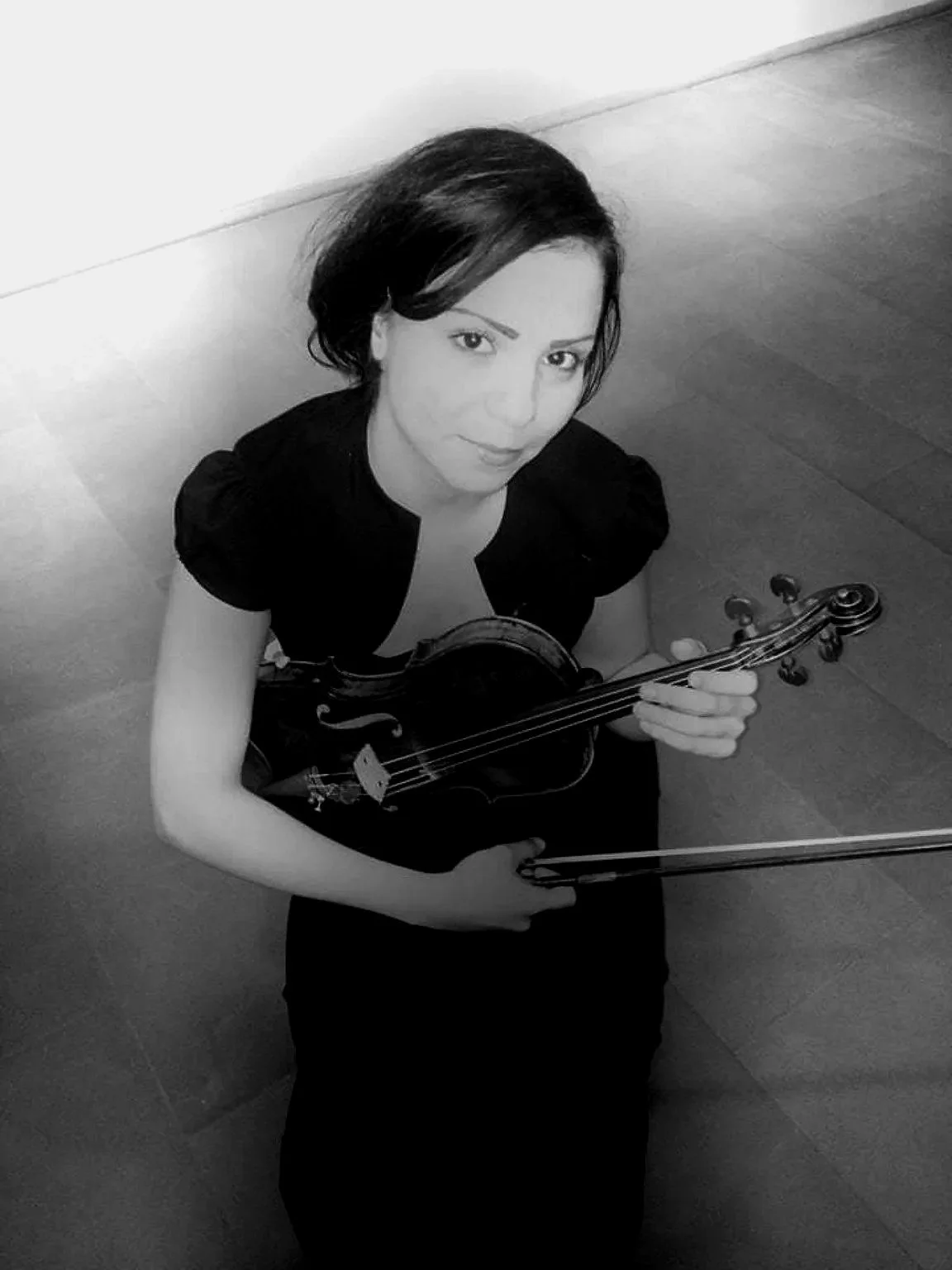 A woman with dark hair sitting on the floor holding a violin and bow, looking up at the camera in black and white.