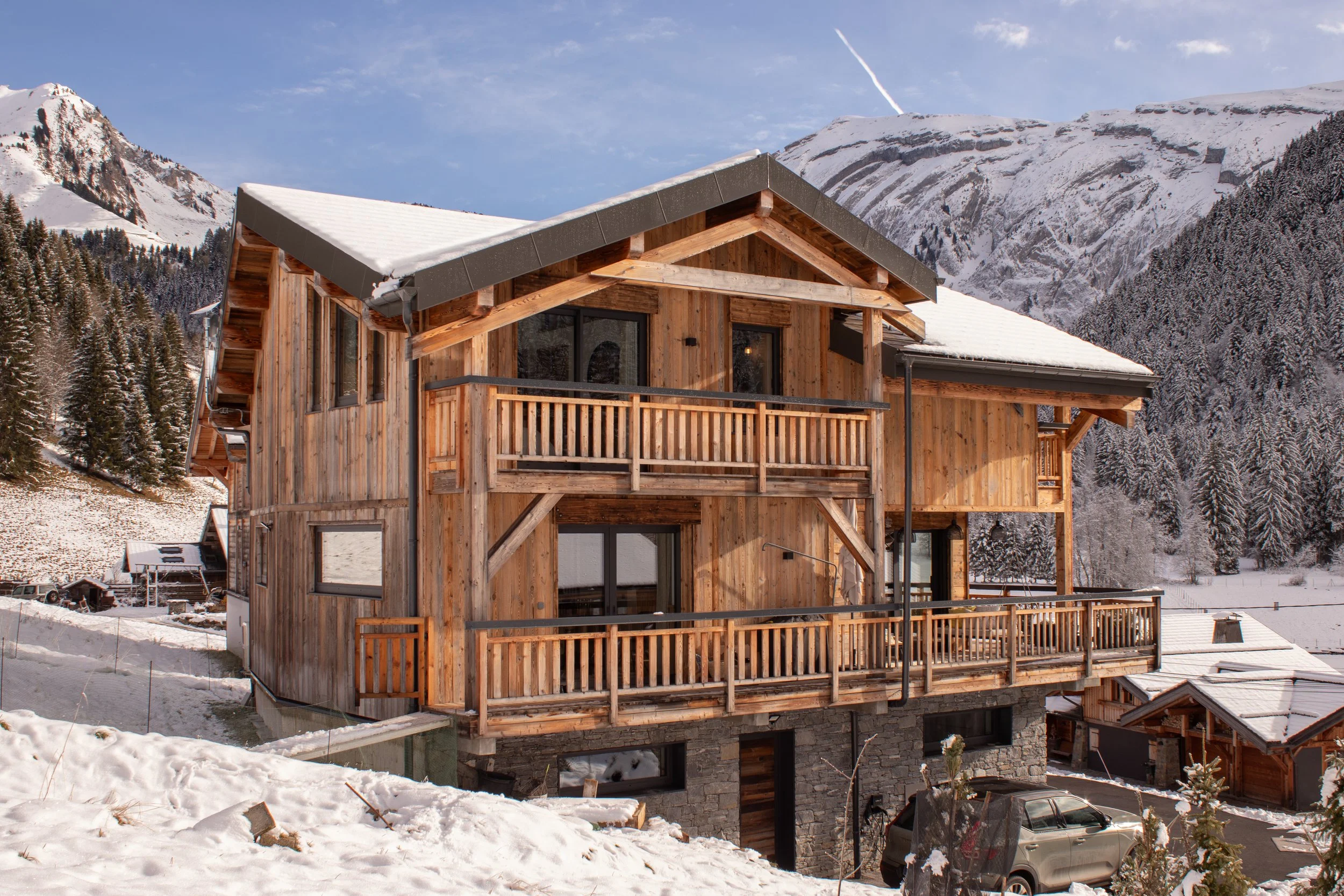 A wooden chalet-style house with multiple balconies, situated in a snowy mountainous landscape, with snow-covered peaks and pine trees in the background.