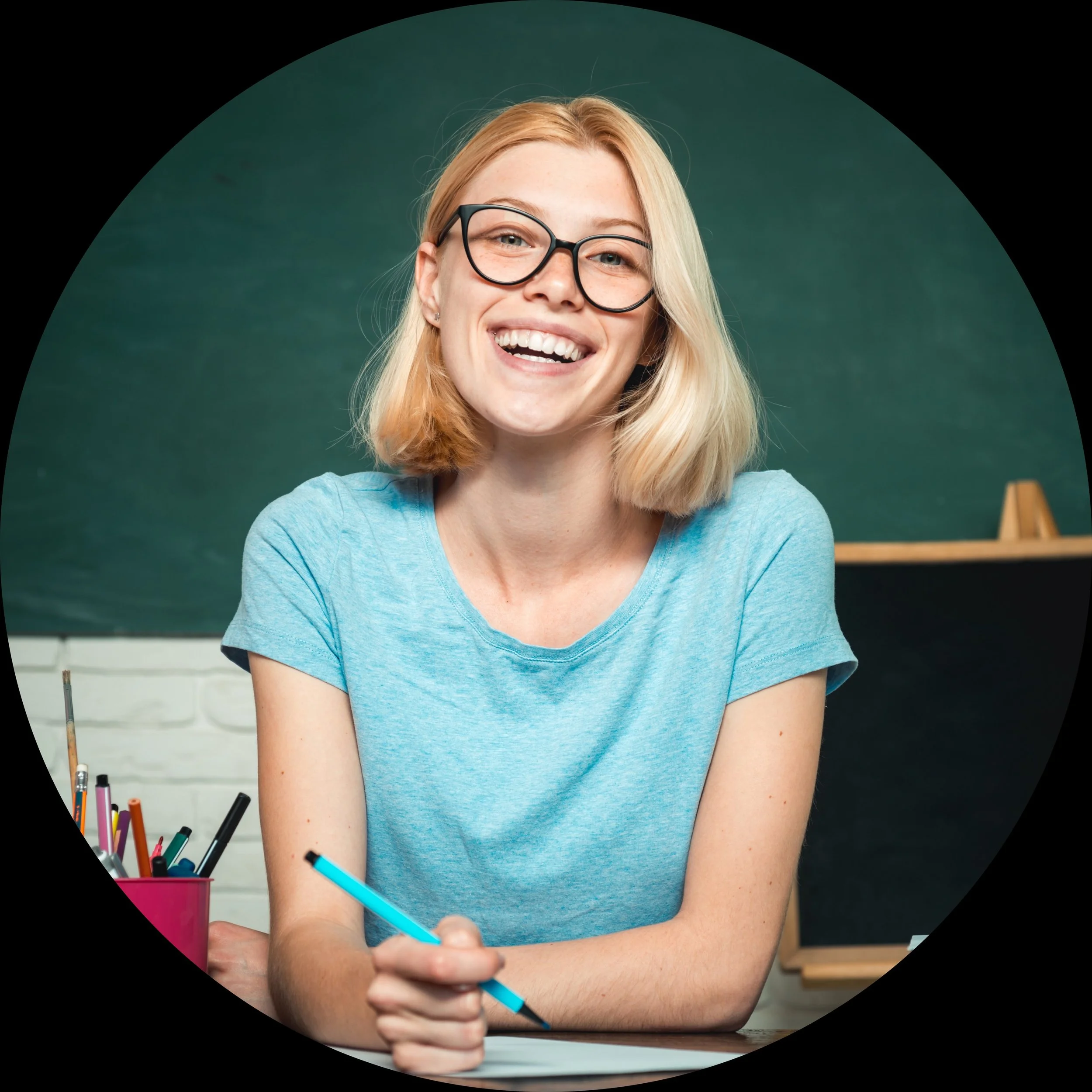 A young woman with glasses and shoulder-length blonde hair smiling and holding a blue pen, sitting at a desk in front of a chalkboard.