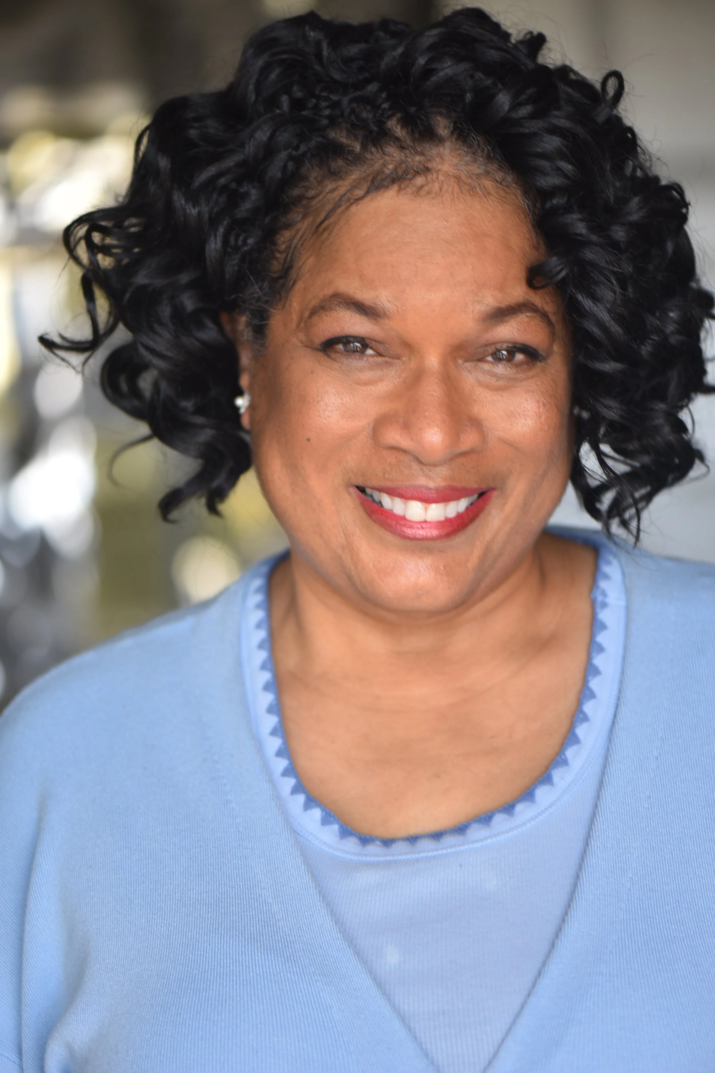 Close-up portrait of a woman with curly black hair, smiling, wearing a light blue top, and standing outdoors.