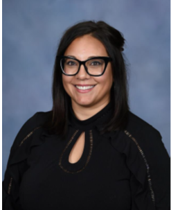 A woman with shoulder-length dark hair, wearing glasses and a black top, smiling against a blue background.