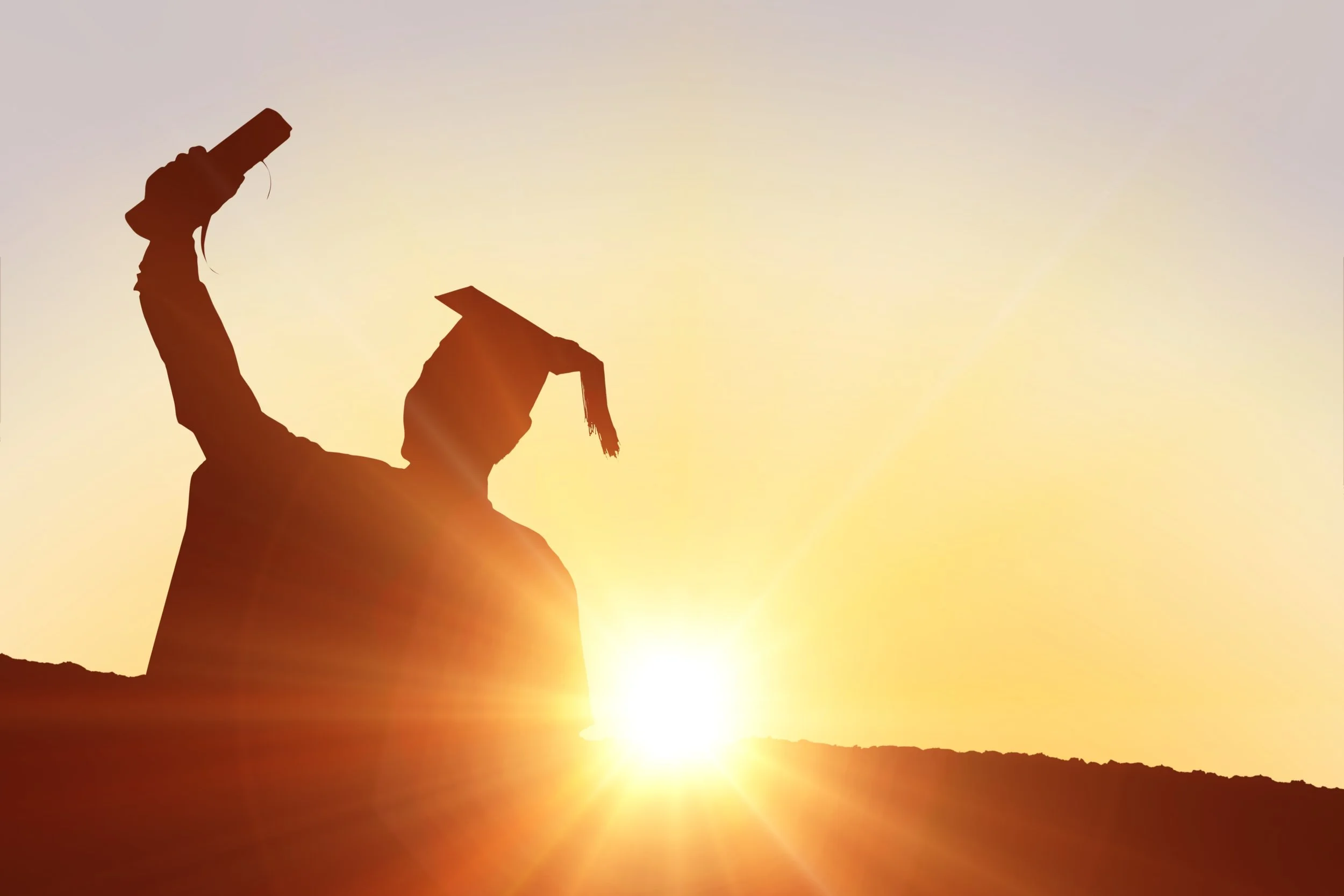 Silhouette of a person holding a phone and wearing a graduation cap, against a sunset or sunrise background.