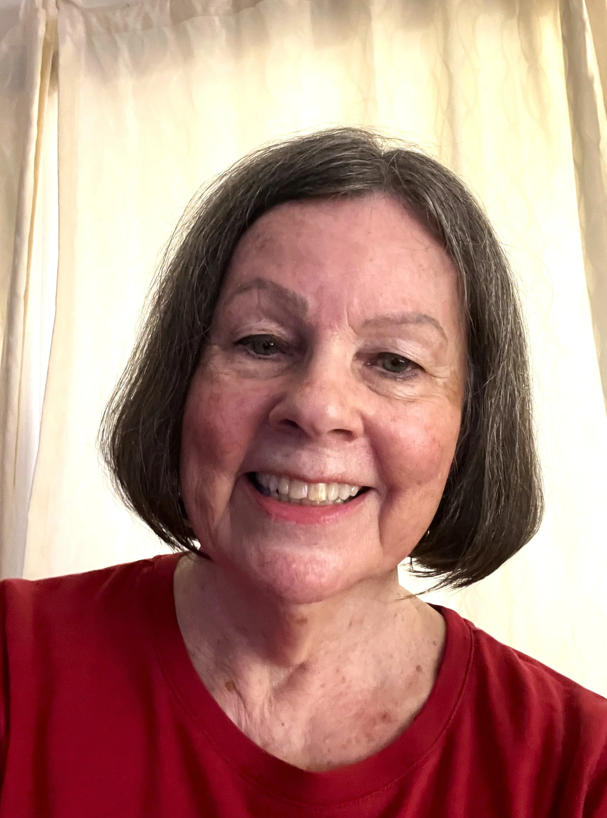 A smiling woman with short brown hair wearing a red shirt standing in front of cream-colored curtains.