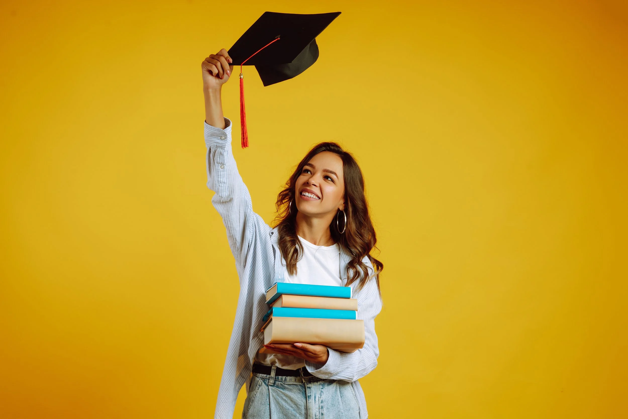 A young woman with long brown hair, wearing hoop earrings, a white shirt, and a light blue striped jacket, holding a stack of books in one hand and a black graduation cap with a red tassel in the other hand, smiling against a yellow background.