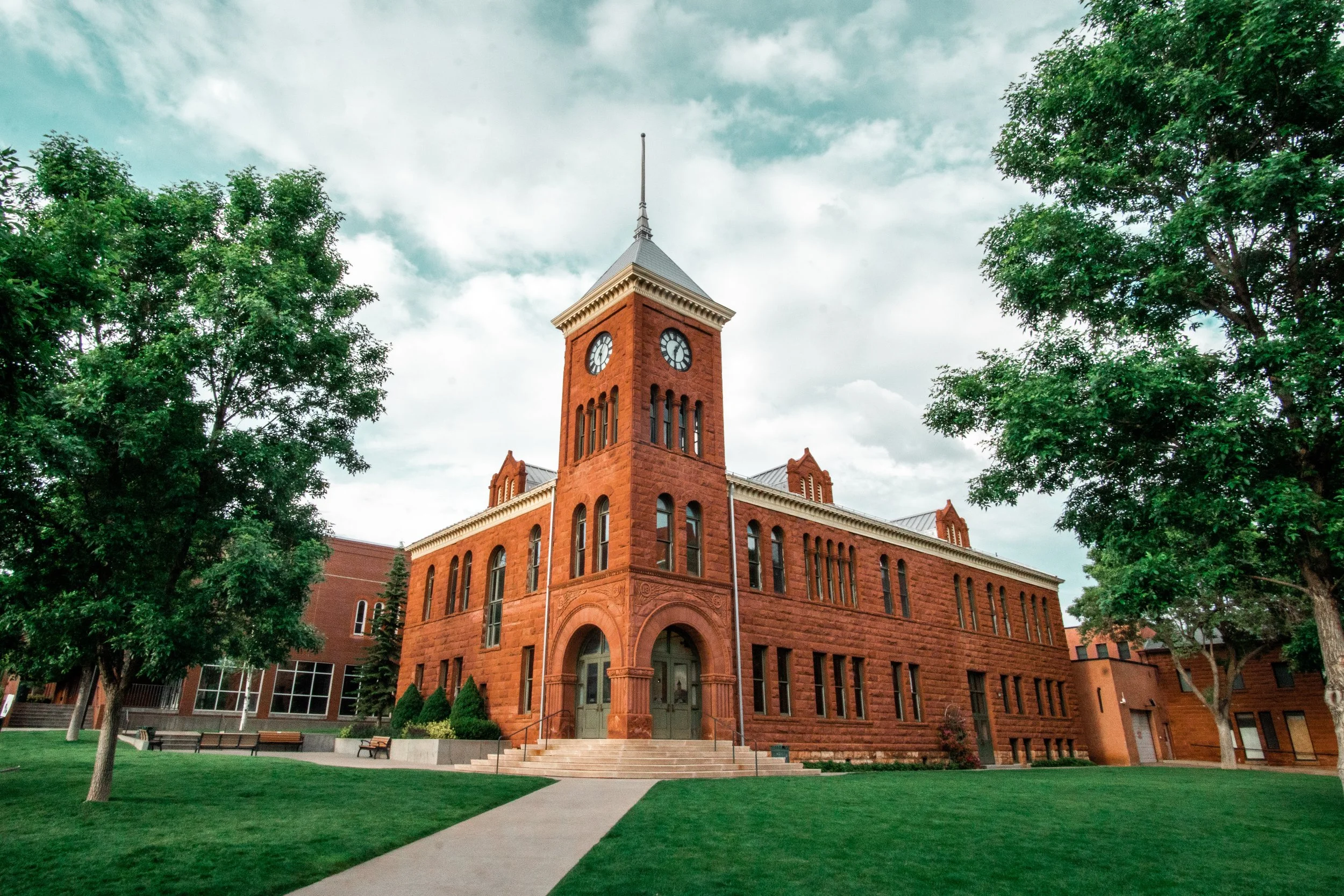 A historic red brick building with a clock tower, surrounded by green trees and a well-maintained lawn.