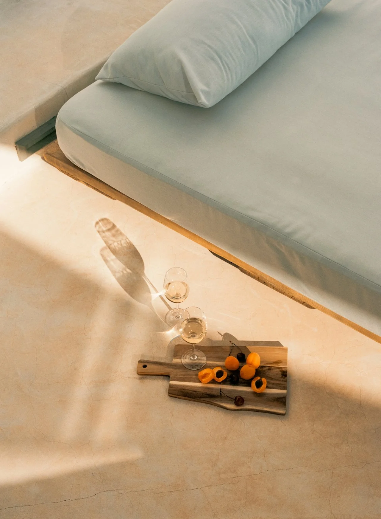 A wooden bed frame with a mattress and pillow, a small wooden serving board with snacks, and two wine glasses filled with white wine on a light-colored floor.