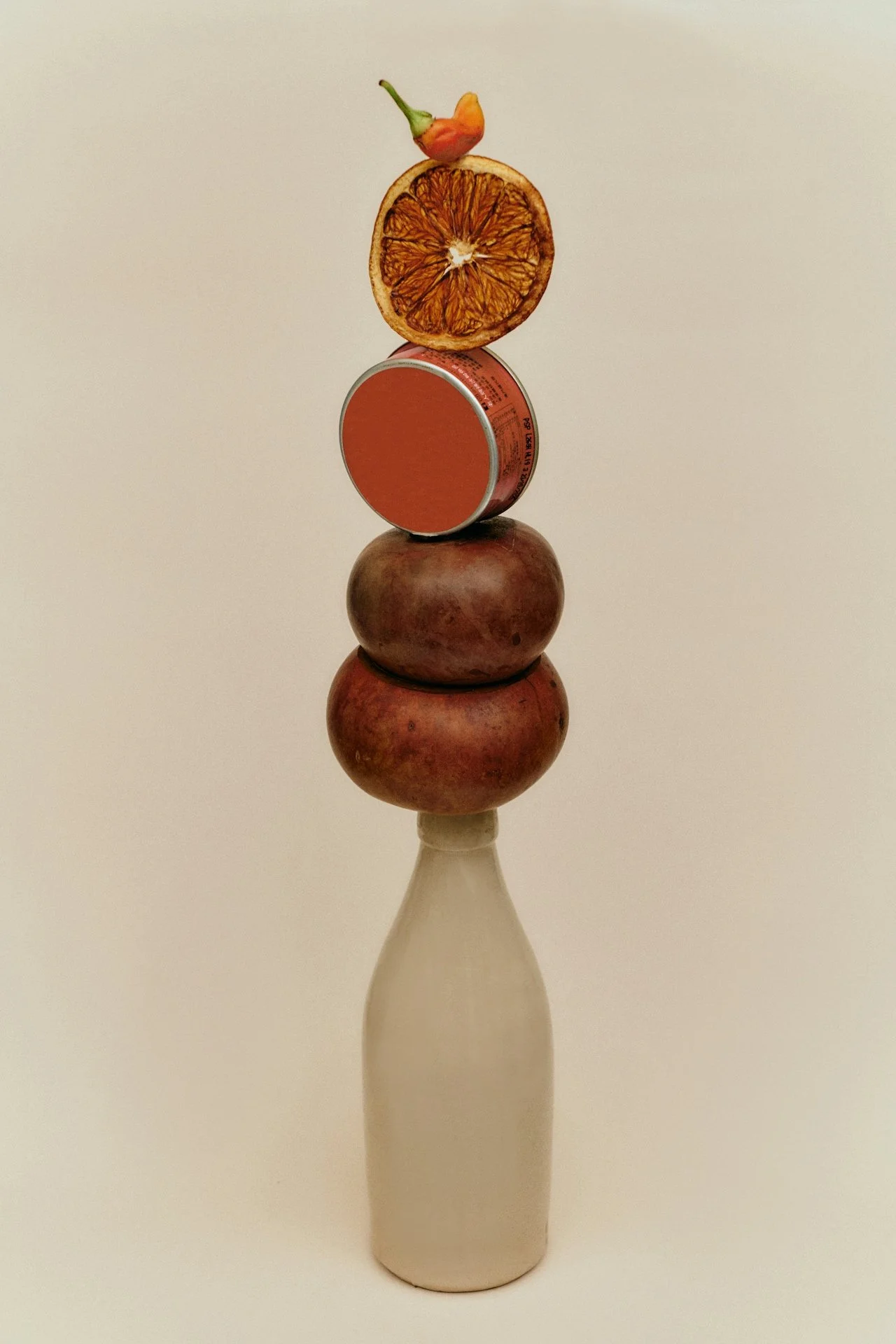 Bottle with two pomegranates, a dried orange slice, a small can, and a small hot pepper stacked on top.