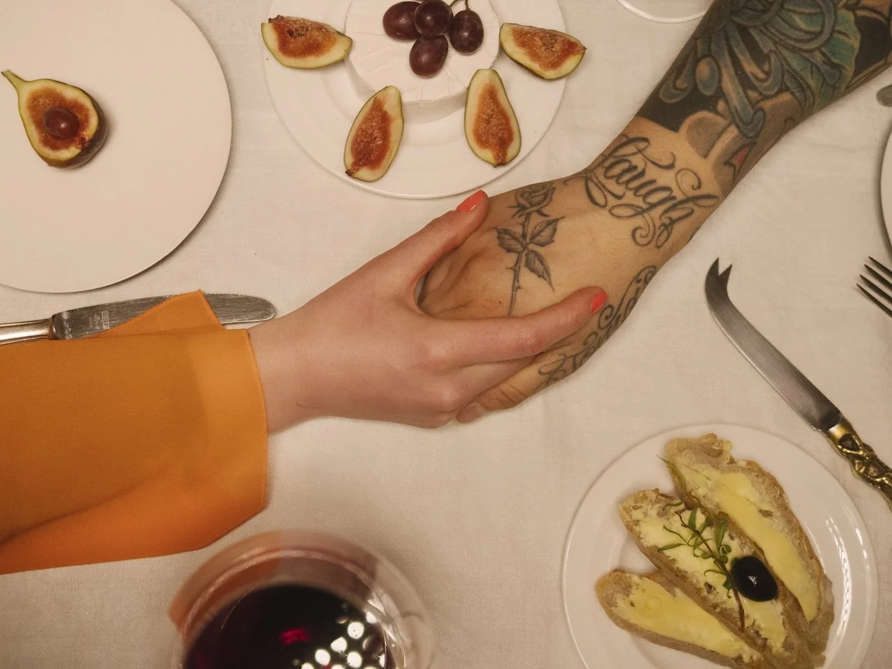 Two people holding hands at a dinner table with plates of figs, grapes, and a slice of cake with yellow filling and a cherry topping, along with a glass of red wine.