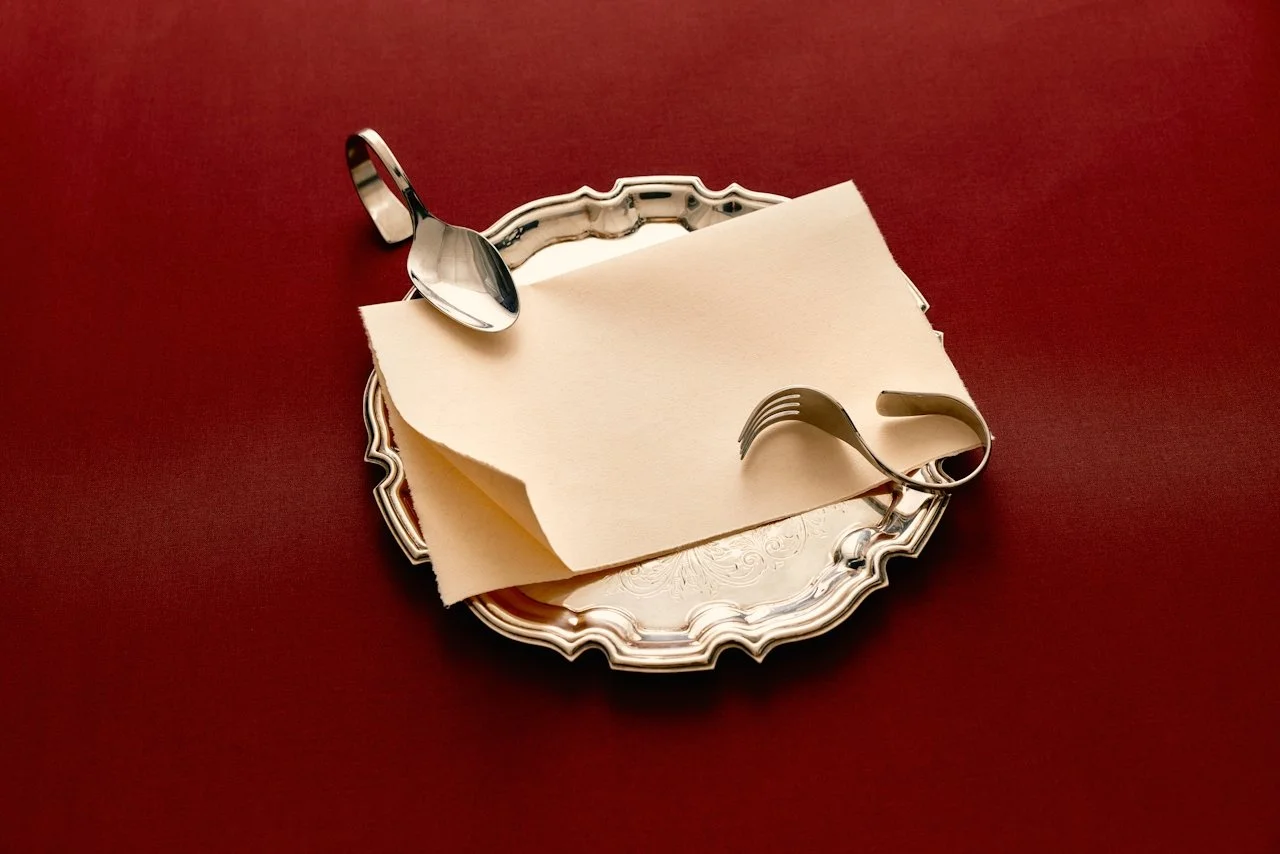 Silver tray with ornate edges, a folded napkin, a spoon, and a fork on a red table.