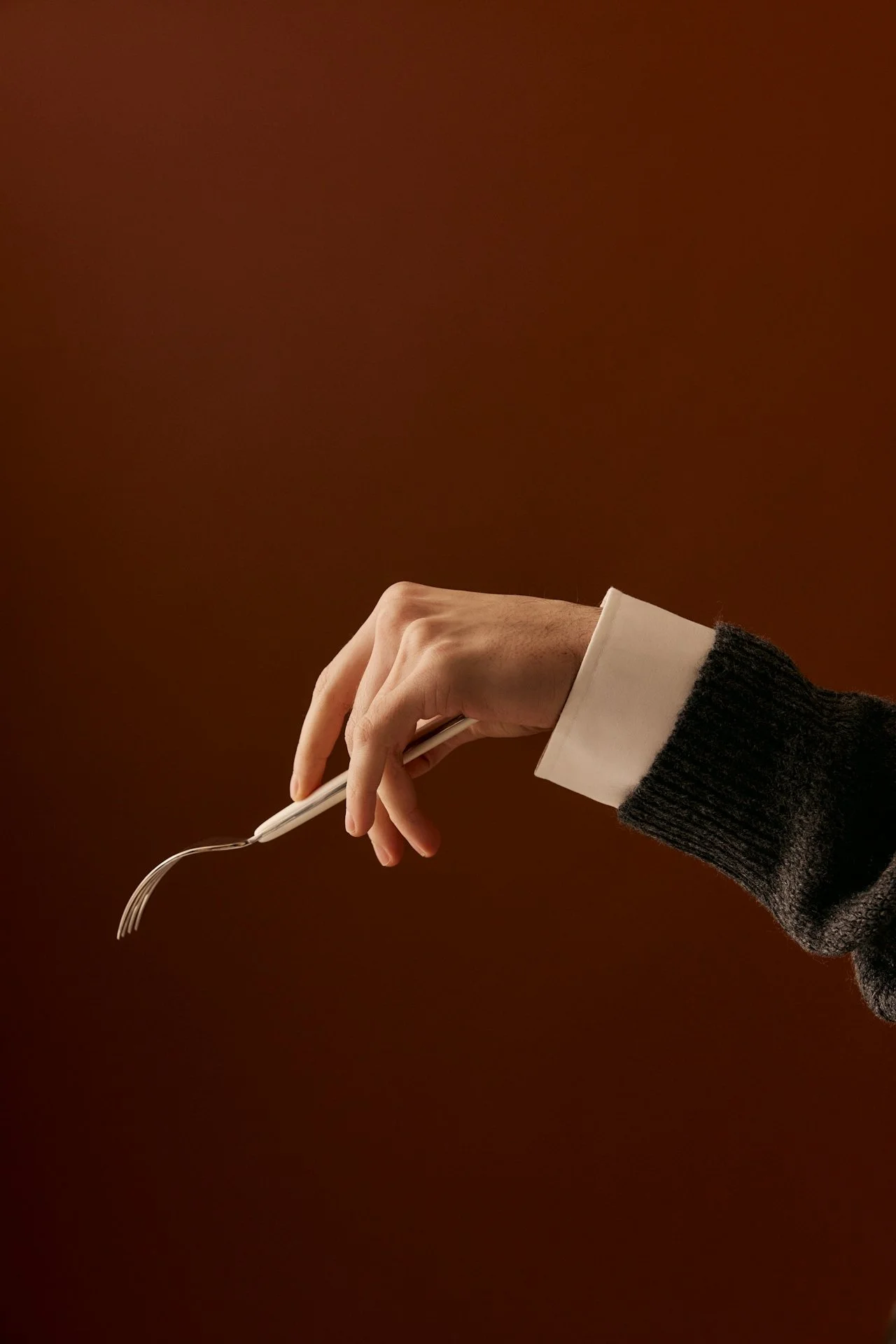 A person's hand holding a white fork against a dark brown background, wearing a dark sweater with a white shirt cuff.