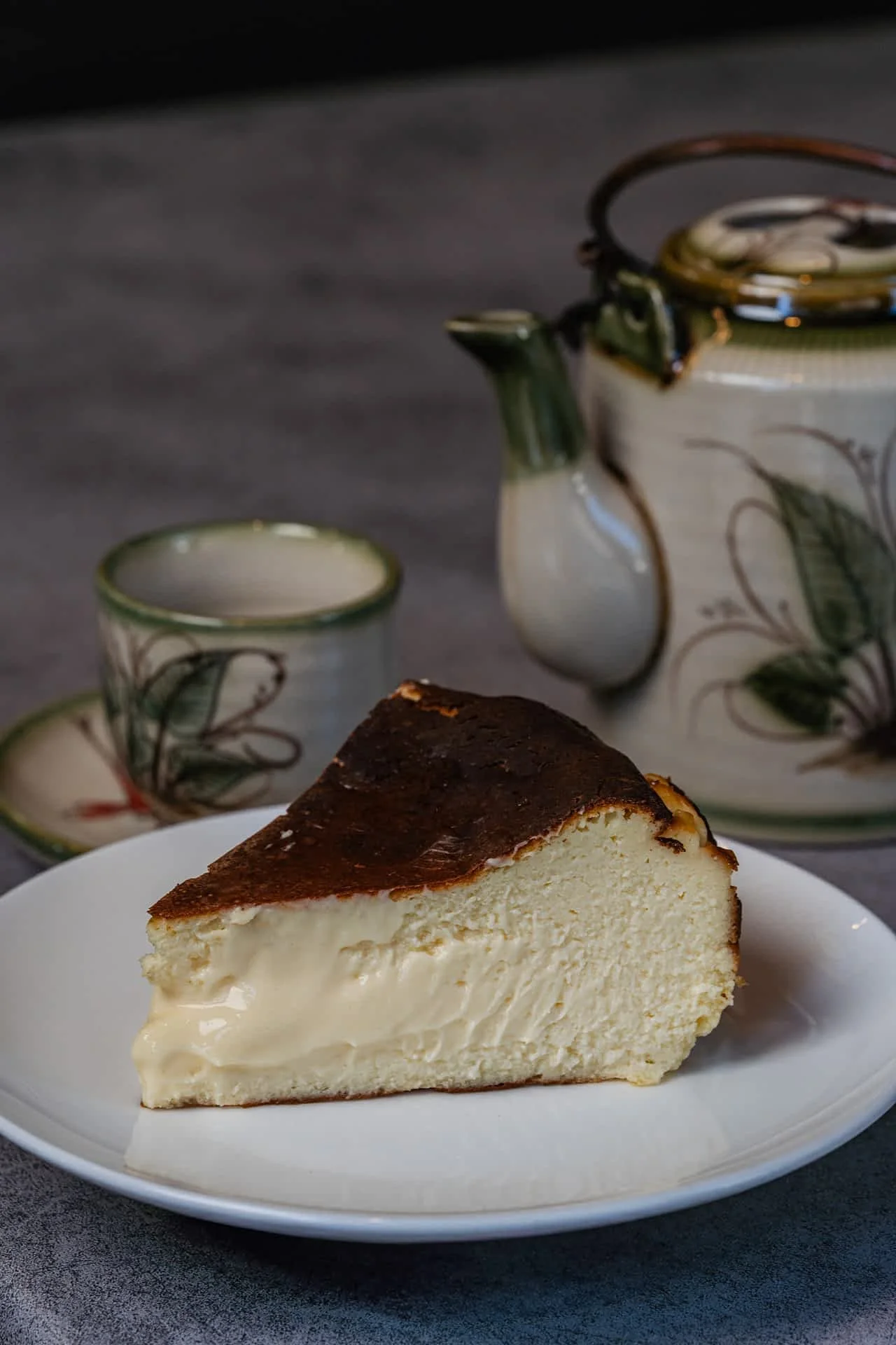 Slice of cheesecake on a white plate with a tea set, including a teapot and a cup, in the background.