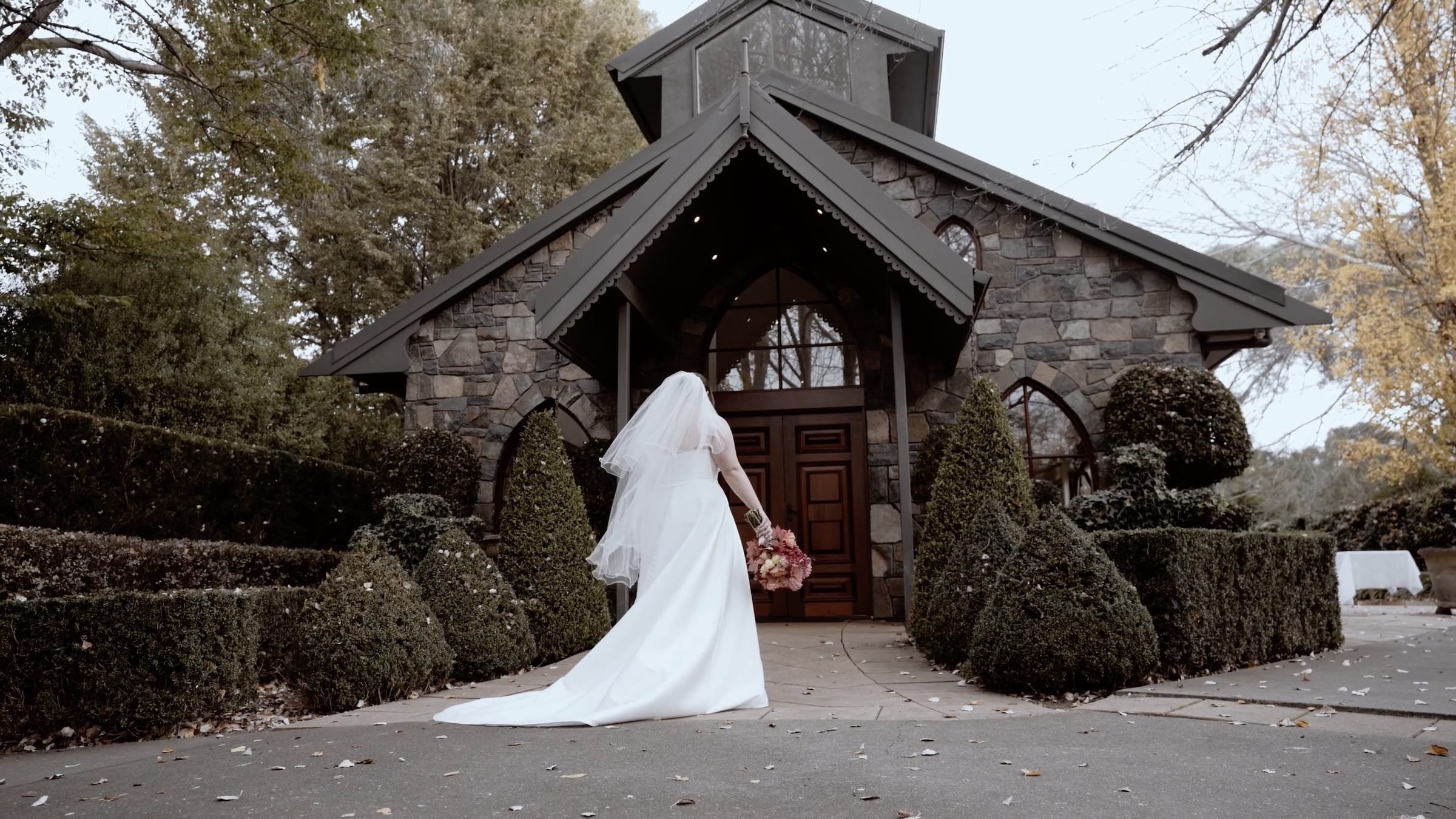 Bride in a white wedding dress holding a bouquet, walking towards a stone church with a peaked roof and glass windows, surrounded by neatly trimmed bushes and trees.