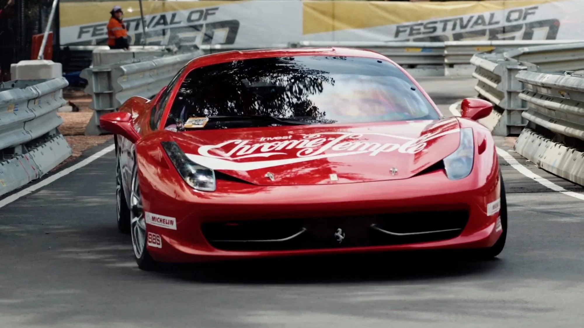 Red Ferrari race car with 'Ferramenta' and other sponsor logos on the hood, driving on a race track with barriers and a person in an orange safety vest in the background.