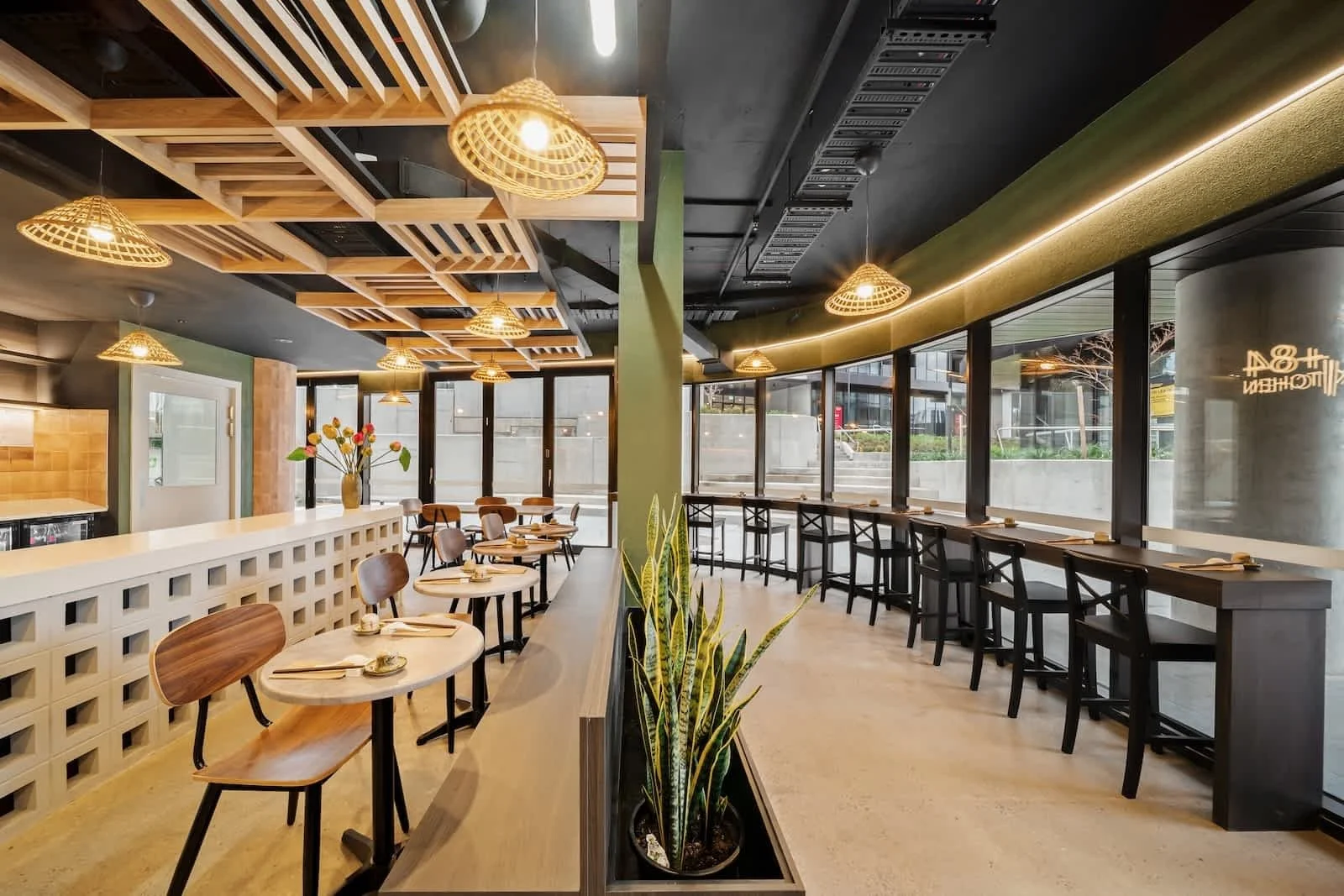 Modern interior of a restaurant or cafe with large windows, pendant bamboo lights, wooden tables and chairs, a plant in a planter, and minimalistic decor.