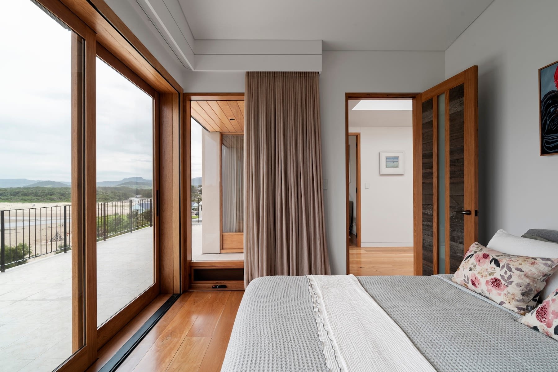 Interior of a bedroom with sliding glass doors leading to a balcony, beige curtains, a bed with white linens and floral pillows, wooden doors, and a hallway with art on the wall.