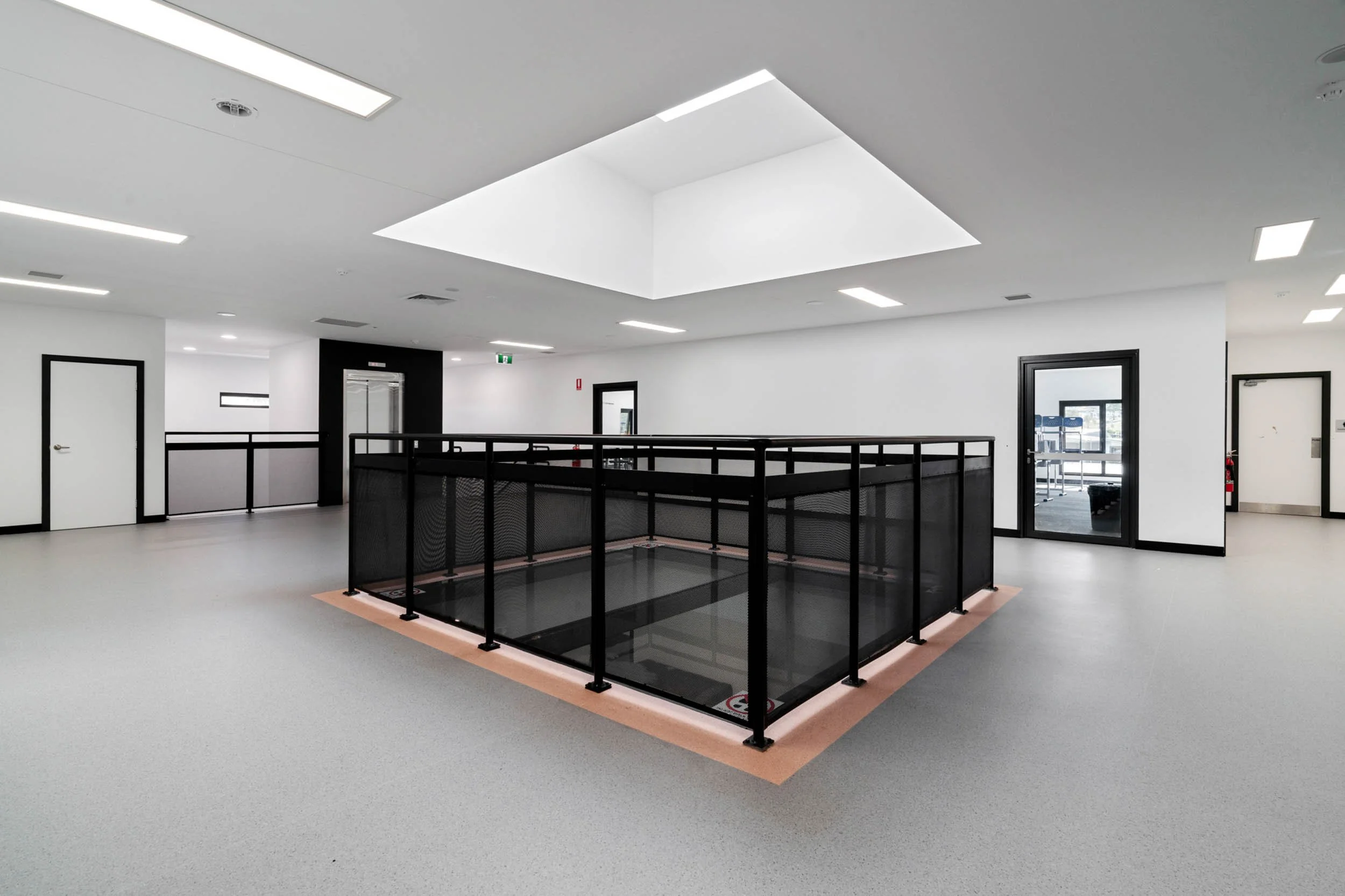 Empty indoor space with grey flooring, white walls, black-trimmed doors, and a black-railed indoor balcony or elevator shaft in the center.