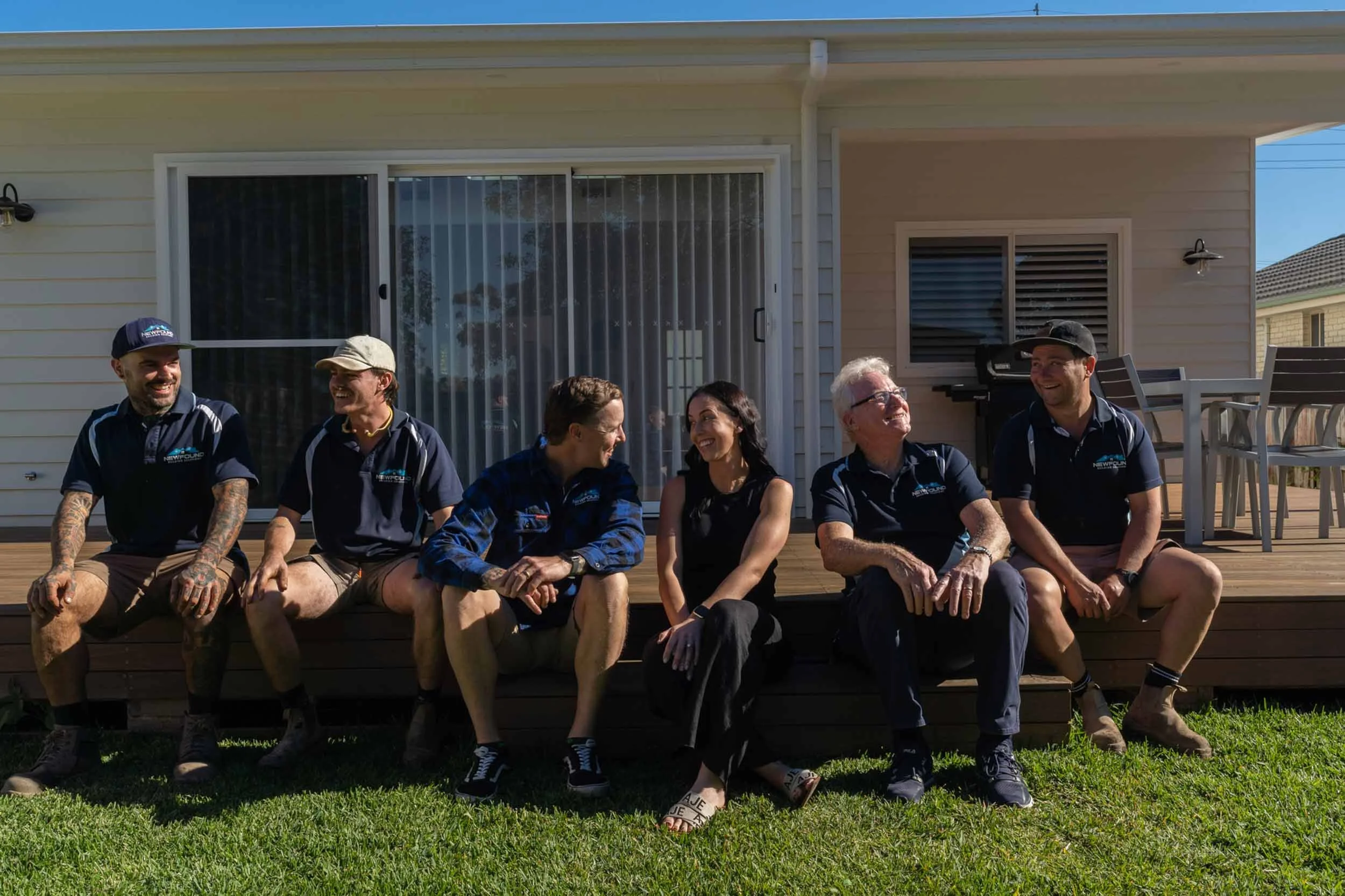 Six people sitting on a wooden porch of a house, smiling and talking to each other. The house has sliding glass doors, windows, and outdoor furniture.