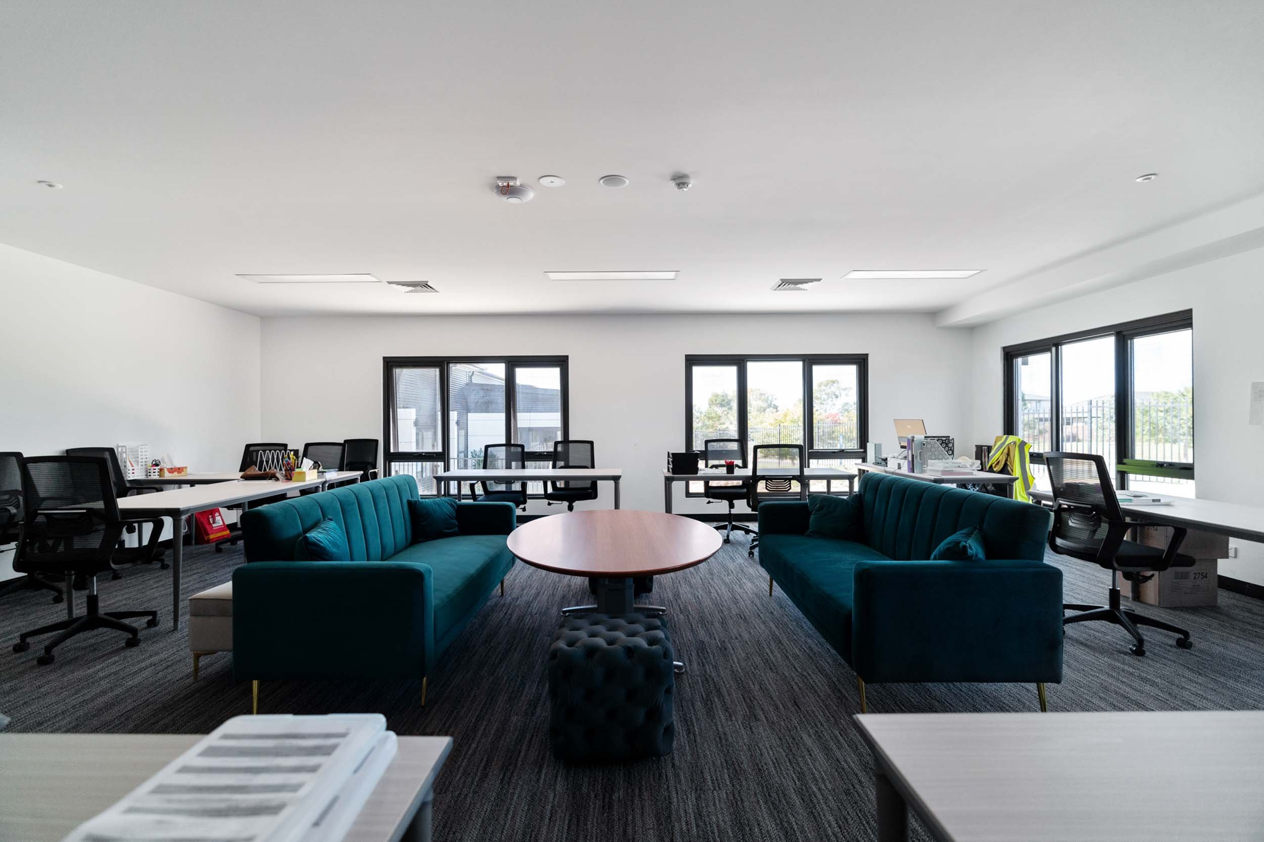 Empty modern office with two teal sofas facing each other, a round wooden table in the center, multiple desks with office chairs on a gray carpet, and large windows bringing in natural light.