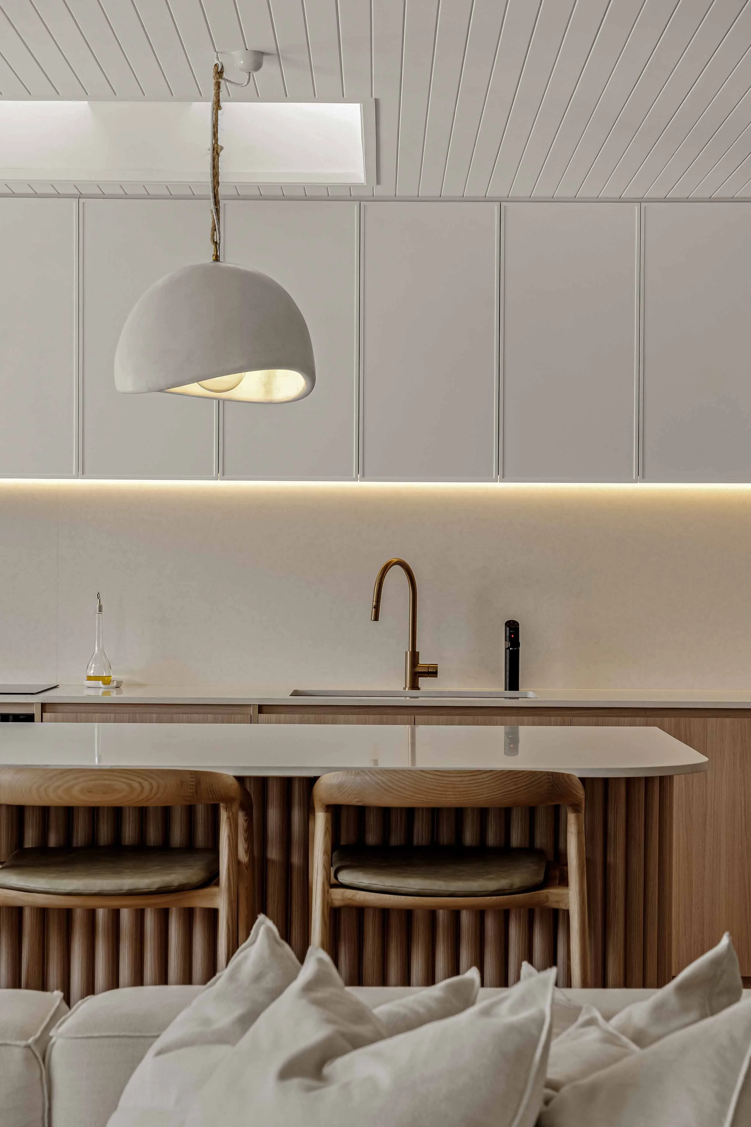 A modern minimalist kitchen with white cabinets, a white countertop, and wooden bar stools with cushions, featuring a brass faucet and a pendant light.