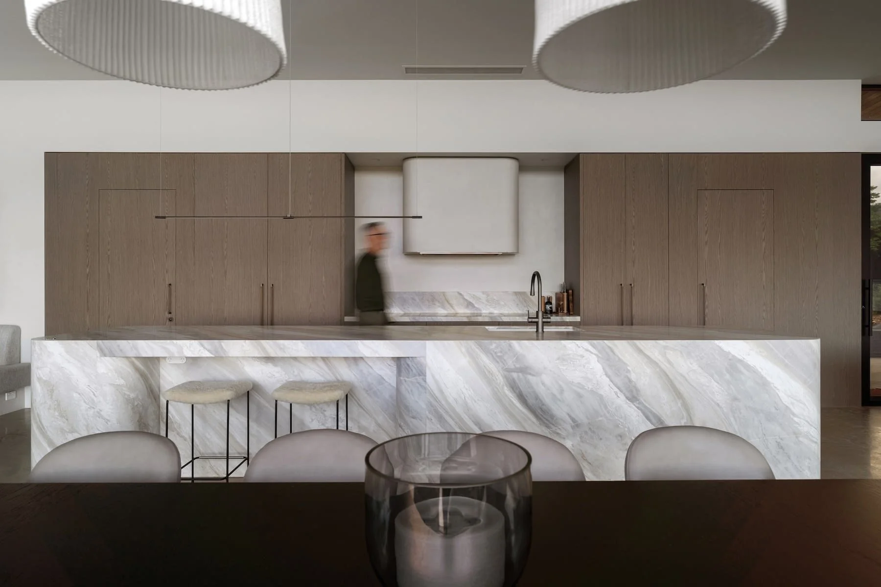 Modern kitchen with a marble island, wooden cabinets, and a blurred man standing behind the island. There are ceiling lights and a candle holder with a candle on a dark table in the foreground.