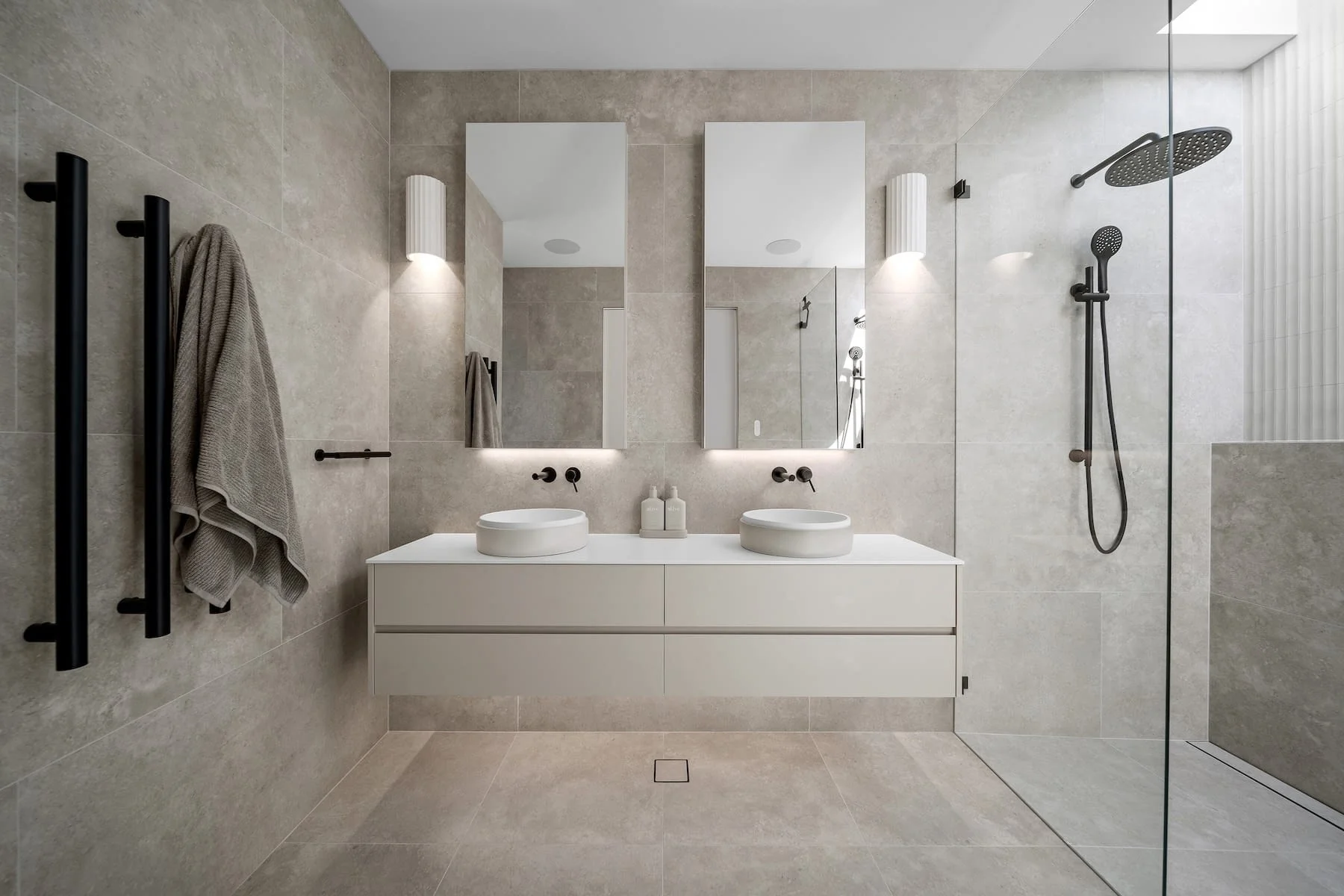 Modern bathroom with beige tiled walls and floor, double vanity with two round vessel sinks, two large mirrors, wall-mounted black faucets, wall sconces, black towel rack with a beige towel, and a walk-in shower with black rain showerhead and handheld shower, enclosed with clear glass.
