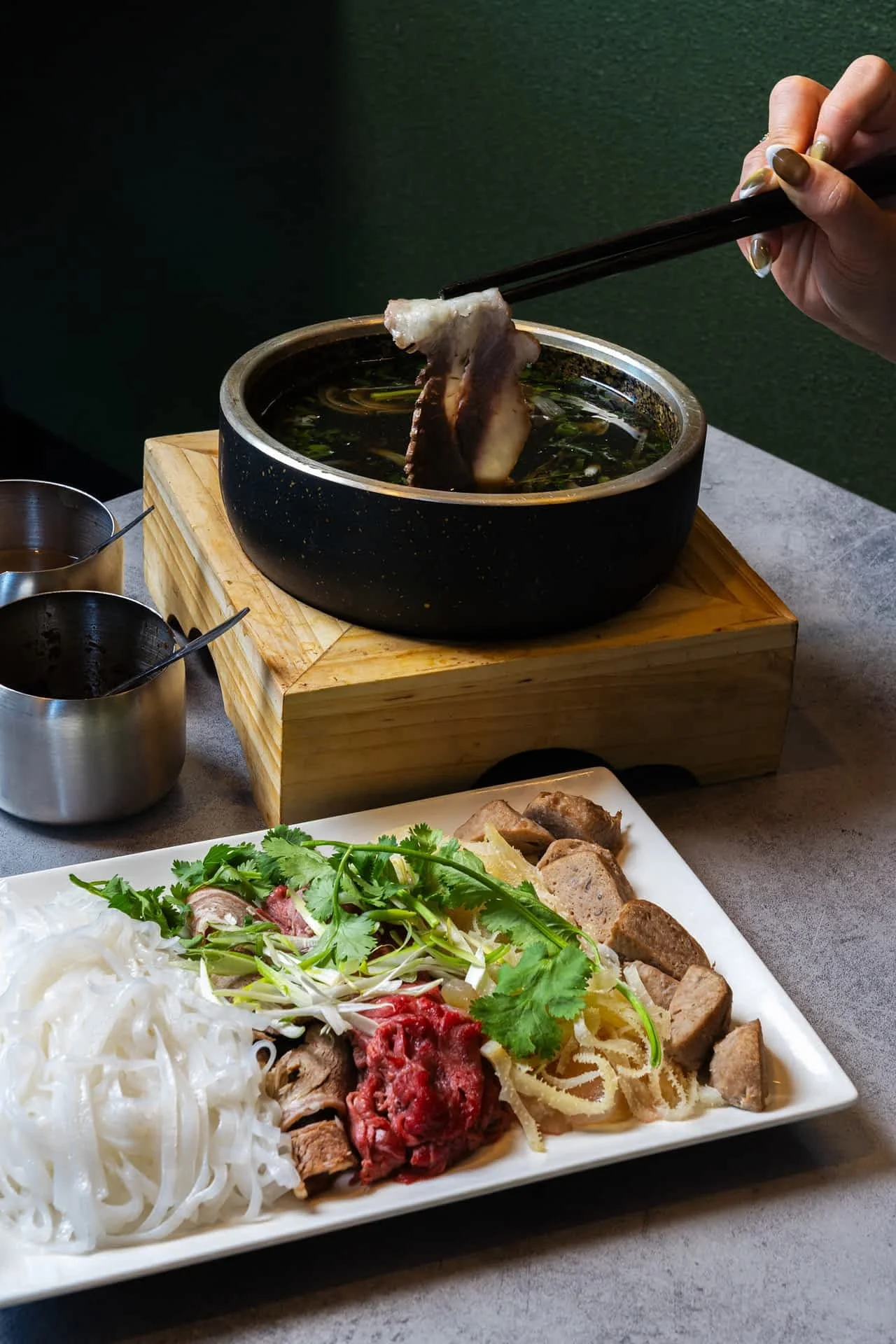 A person using chopsticks to dip a piece of meet into a hot pot with broth, with a plate of sliced meat, herbs, noodles, and vegetables in the foreground.