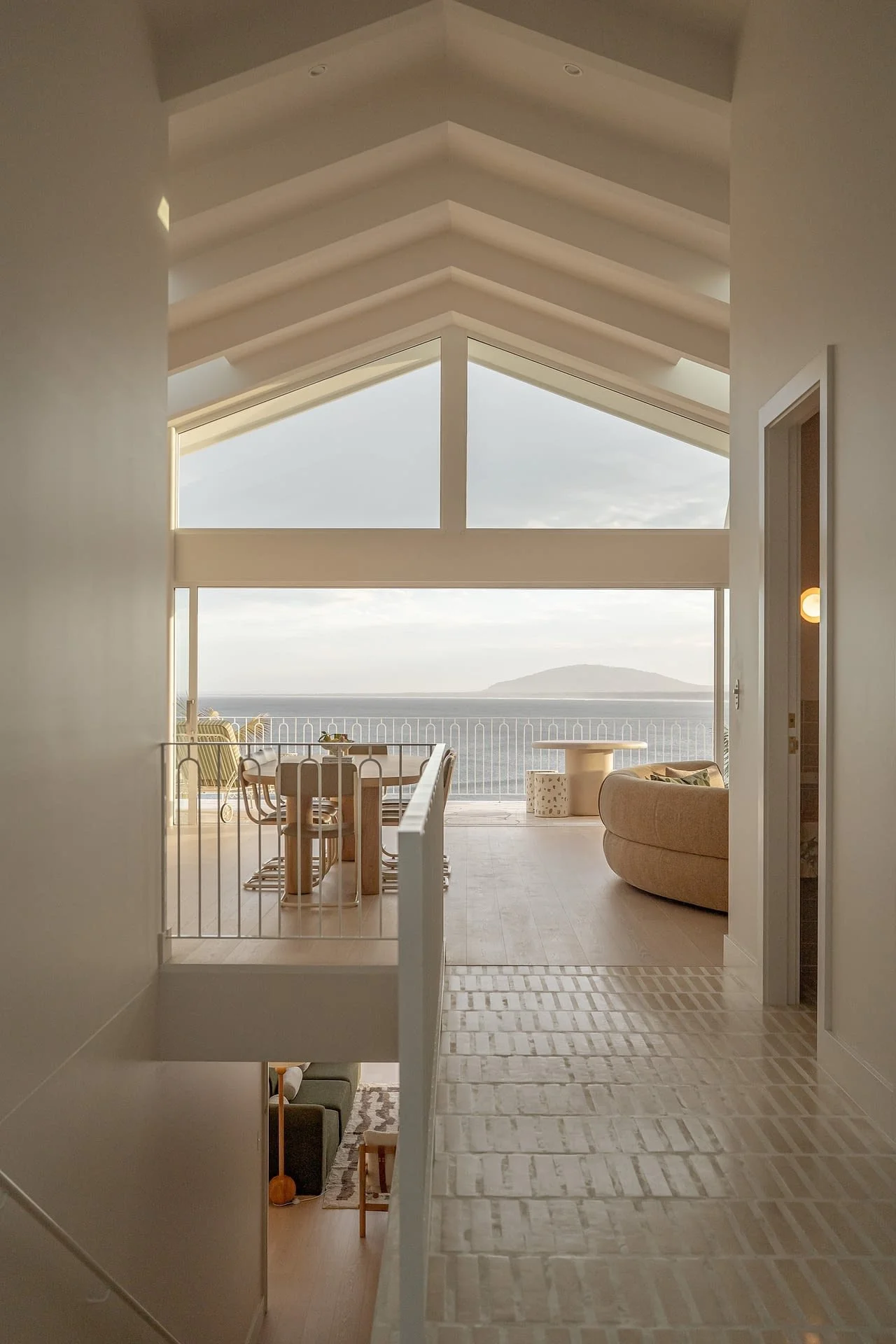 Interior view of a modern home with a spacious room overlooking the ocean through large glass windows, featuring a dining area with a table and chairs, a curved sofa, and outdoor furniture on a balcony.