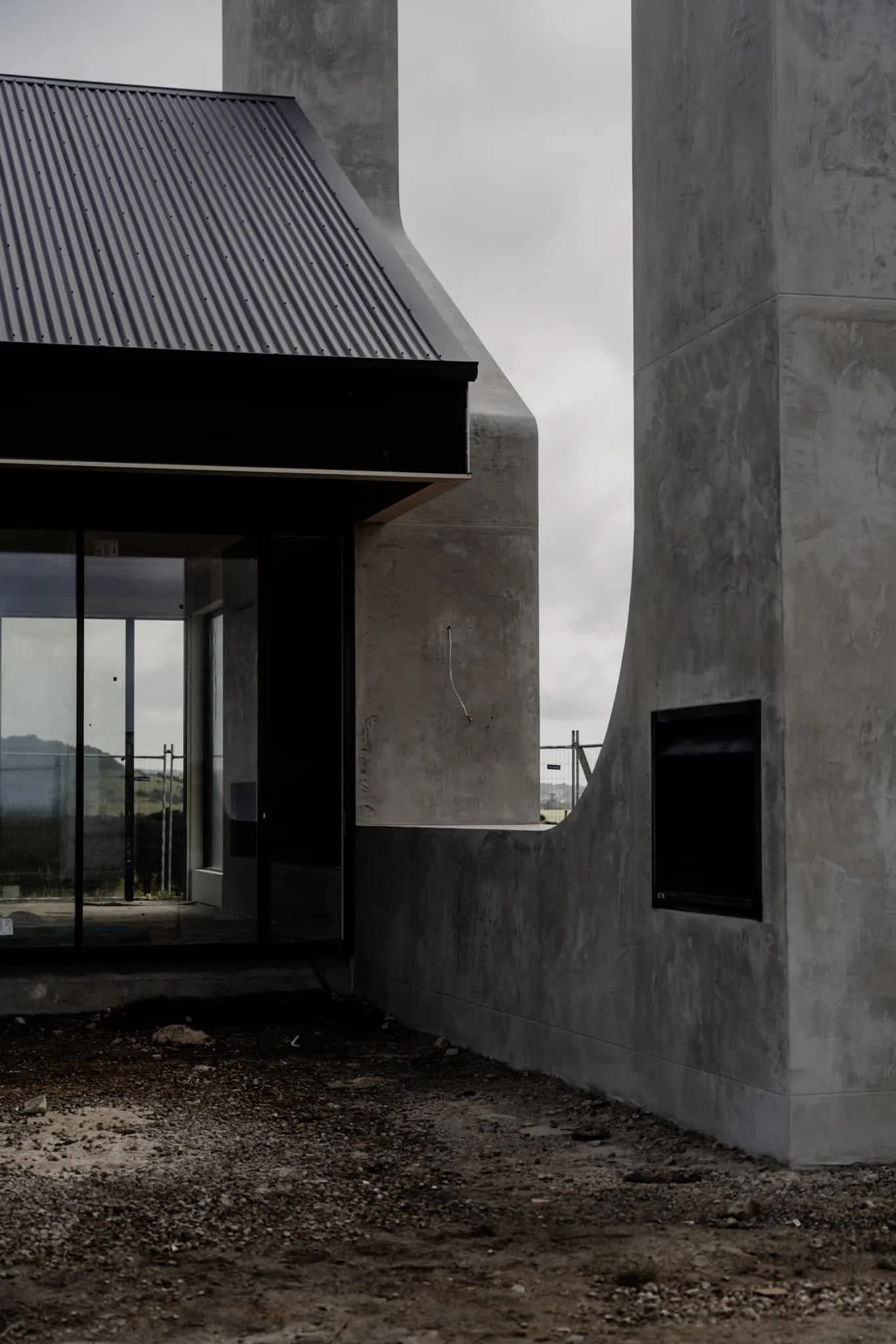 Close-up of a modern building under construction, featuring concrete walls, large glass doors, and a sloped metal roof, with a cloudy sky in the background.