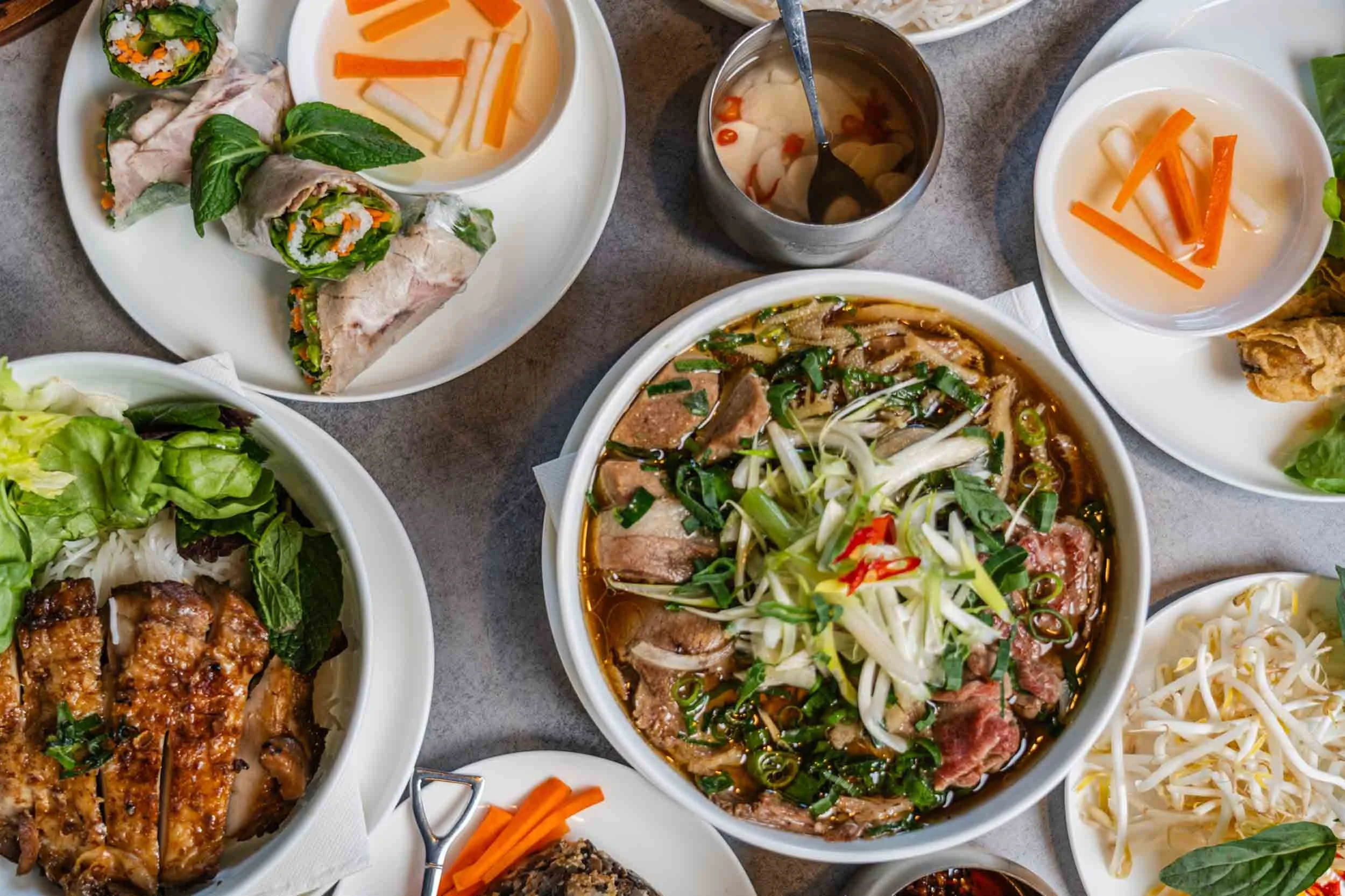 A variety of Vietnamese dishes on a table, including pho soup with beef and herbs, spring rolls, grilled chicken, fresh vegetables, and side dishes.