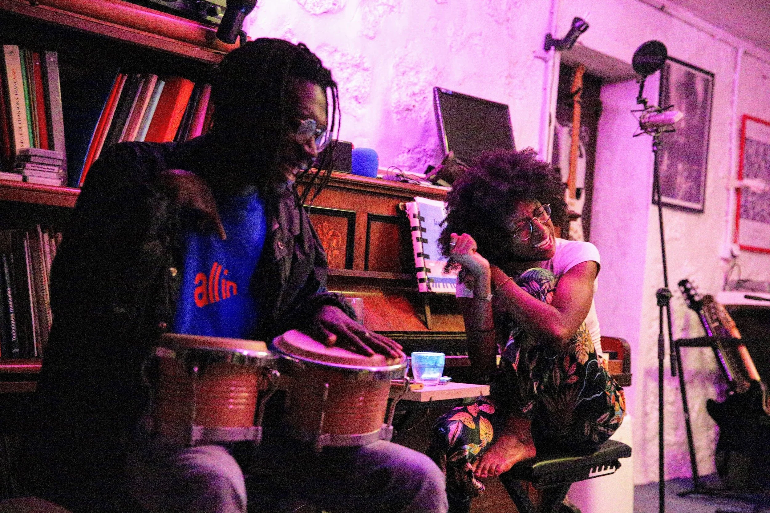 Two people at an indoor music session: one person playing bongos and the other sitting on a chair, smiling and resting their hand on their head. The room features a bookshelf, a piano, and music equipment, with purple lighting.