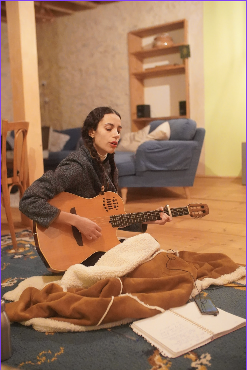 A woman with dark hair and braids is sitting on a rug, playing an acoustic guitar. She is in a cozy room with a blue sofa, a wooden shelf, and a stone wall in the background.