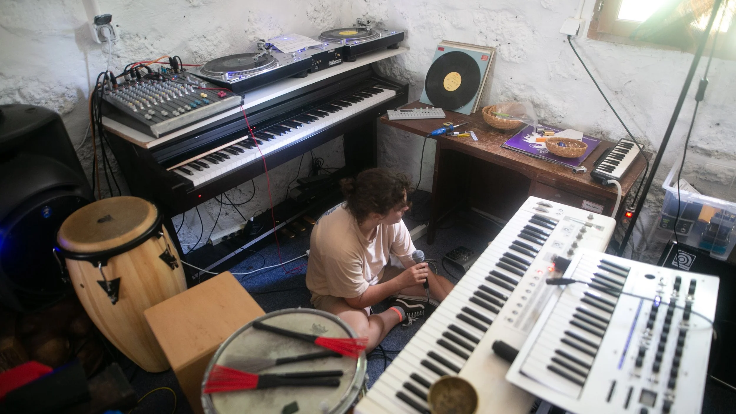 A young person sitting on the floor holding a microphone surrounded by musical instruments and equipment in a cluttered room with white brick walls.