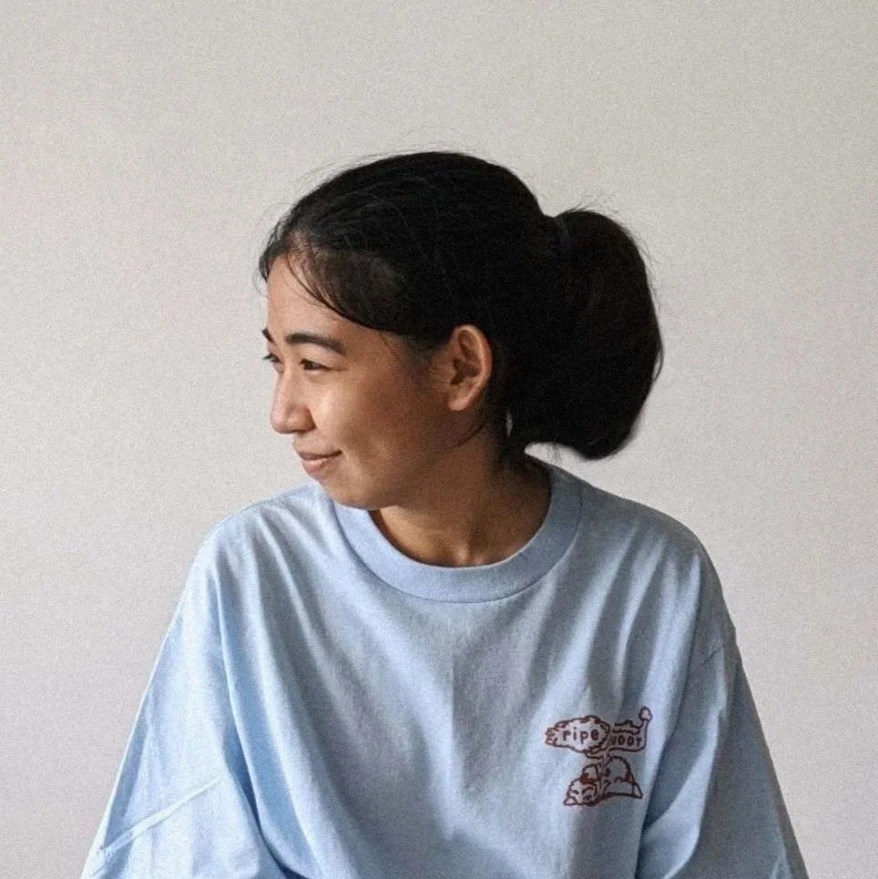 A young woman with black hair tied in a ponytail, smiling and looking to the side, wearing a light blue t-shirt with a small graphic on the chest, against a plain white background.