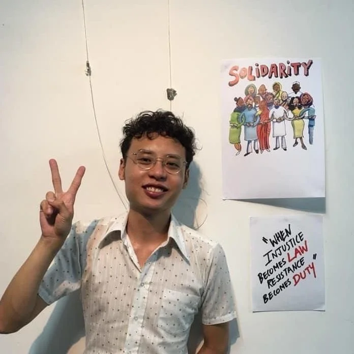 Young man with curly hair and glasses making a peace sign next to a poster that says "Solidarity" and another note with a quote about justice, law, resistance, and duty.