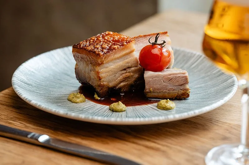 Ein Teller mit einem Stück Schweinebauch, einem kleinen Tomaten, einem Klecks Senf und einer dunklen Soße, daneben ein Glas Bier.