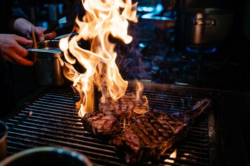 Steak auf dem Lavastein Grill mit offenen Flammen, Chef benutzt Zange im Hintergrund