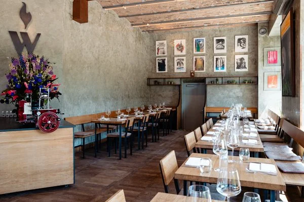 Ein stilvoller Restaurantraum der Weingrün Rotisserie mit Holztischen, Stühlen, Wein- und Wasser glasses, einer Wanddeko aus mehreren Kunstwerken und einer Blumenarrangement auf einem Sideboard.