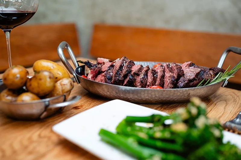 Rinderfilet in einer Metallservierschale, Kartoffeln in einer Schale, grüner Spargel auf einem weißen Teller, Glas Rotwein im Hintergrund.