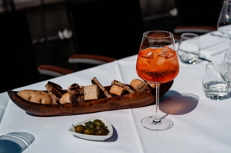 Ein gedeckter Tisch mit einer Aperitifflasche, einem Glas mit rotem Getränk und Eis, einem Holztablett mit ofenfrischem Rosmarinbrot, einer kleinen Schale mit grünen Oliven und weiteren leeren Gläsern.