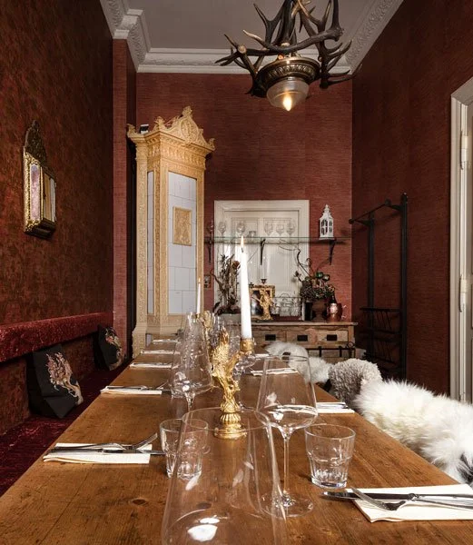 Ein eleganter Essraum im Restaurant Jolesch mit einem langen Holztisch, gedeckt mit Gläser, Besteck und Kerzen. Die Wände sind rot mit Dekorationen, und es gibt einen antiken weißen Kamin im Hintergrund.