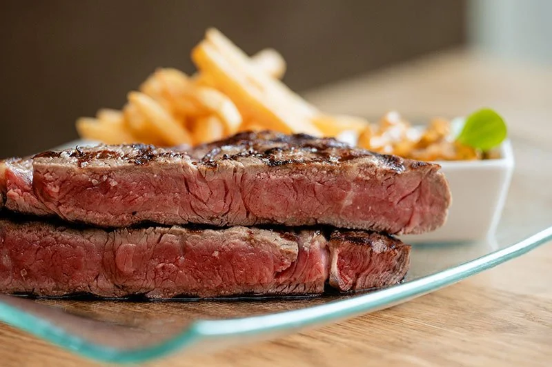 Gegrilltes Steak mit Pommes frites und einer Beilage in einer weißen Schüssel, auf einem Glas- und Holztisch