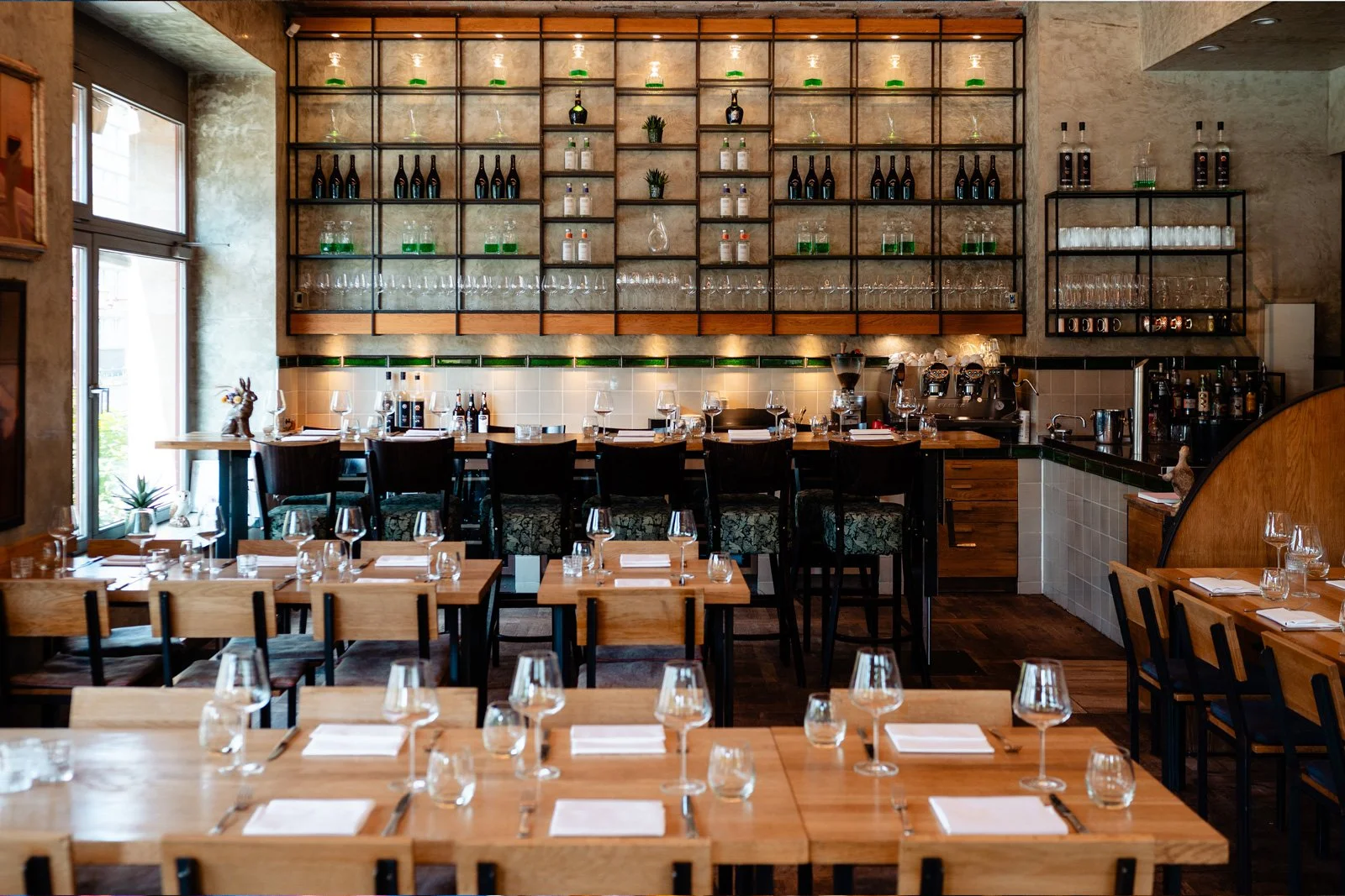 Rotisserie Weingrün, ein stilvolles Restaurant mit Holztischen, Stühlen, Wein- und Cocktailgläsern, einer Wand mit offenen Regalen voller Flaschen, Gläser und Dekorationen, und einem Barbereich im Hintergrund.