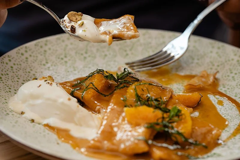Ein Teller mit einem Stück Kürbis, Sauce und einer Portion Sour Cream, garniert mit Kräutern. Jemand hebt ein Stück Kürbis mit einem Löffel. Ein zweiter Löffel steht auf dem Teller.