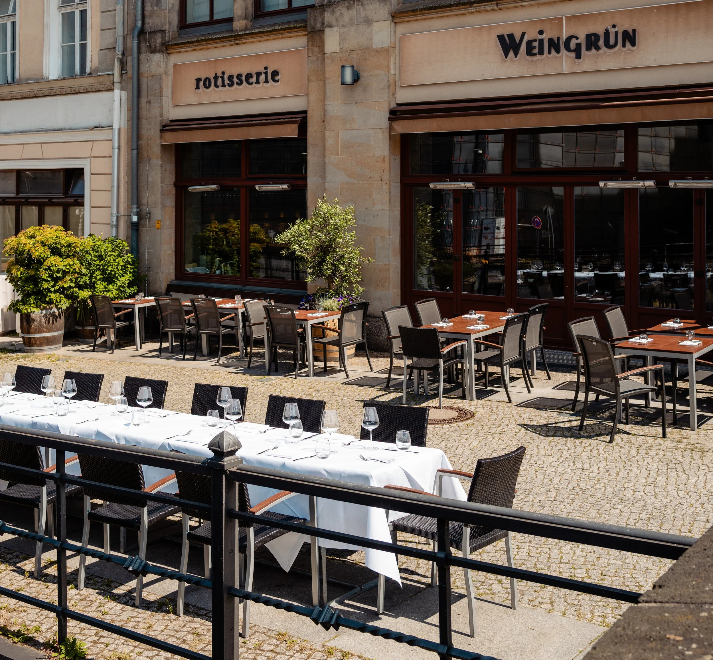 Außenbereich eines Restaurants Weingrün Rotisserie mit weißen und braunen Tischen, umgeben von schwarzen Stühlen, mit Tafelservice und Gläsern, im Hintergrund eine Fassade mit Fenstern und Begrünung, Beschriftungen 'rotisserie' und 'Weingrün'