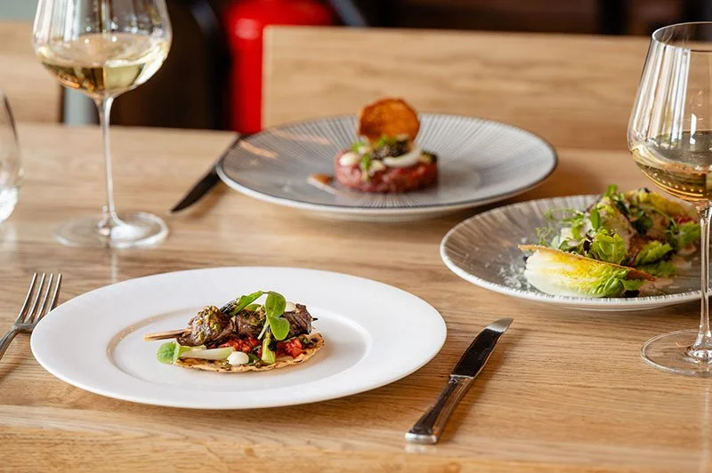 Eleganter Restauranttisch mit drei Tellern, zwei Gläser Weißwein, und Besteck, serviert mit verschiedenen Gourmetgerichten.