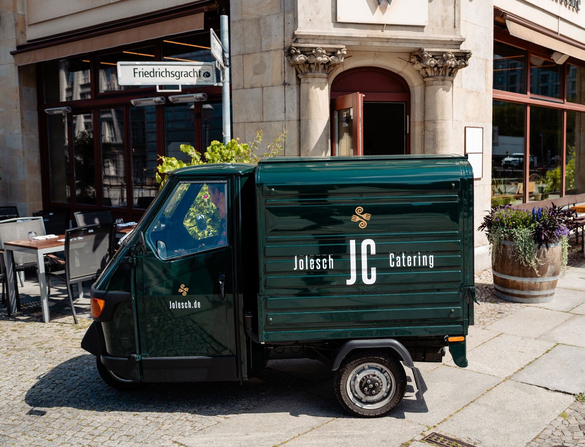 Ape 50 , kleines grünes Lieferfahrzeug für JC Jolesch Catering, das an einer Straßenecke parkt, mit einer blumenbedeckten Straße und dem Restaurant Weingrün Rotisserie im Hintergrund.