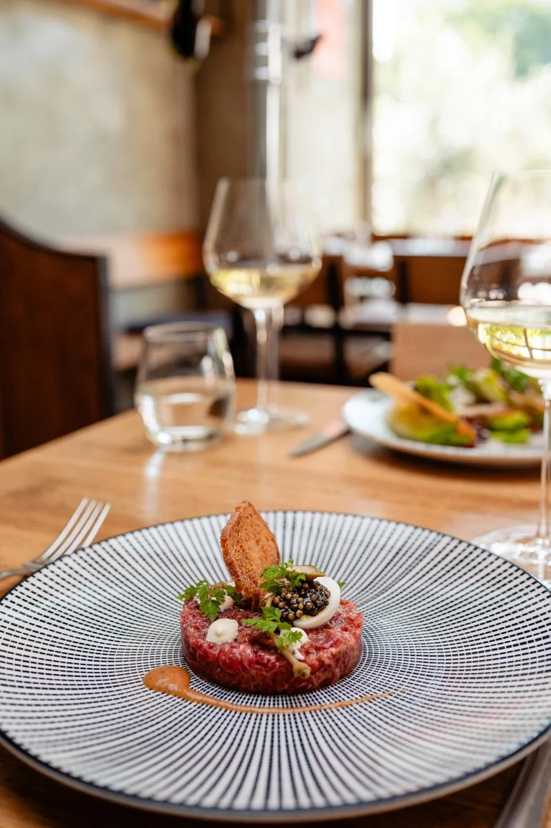 Eleganter Teller mit Tatar vom Weideochsen garniert mit Senf, krossem Knusperbrot, frischen Kräutern und Saucen auf einem Esstisch bei gutem Licht.
