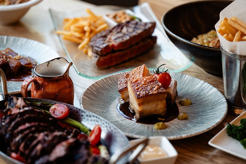 Verschiedene Gerichte und krosser Duroc Schweinebauch auf einem Tisch, darunter Fleisch, Gemüse, Kuchen und Pommes Frites.