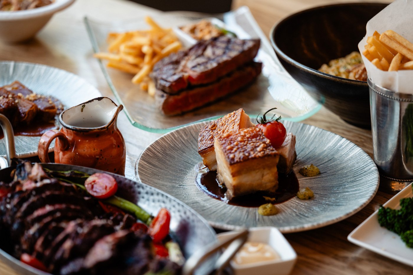 Verschiedene Gerichte auf einem Tisch, darunter gebratenes Fleisch, Krosser Duroc Schweinebauch, Pommes frites, Gemüse, und Dips.